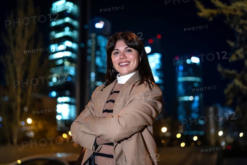 Young professional woman smiling at camera, crossing arms, standing confidently in an urban environment at night with blurred city skyscrapers in the background