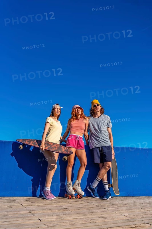 Three cool skaters enjoying sunny day, posing with skateboards and roller skates against vibrant blue wall