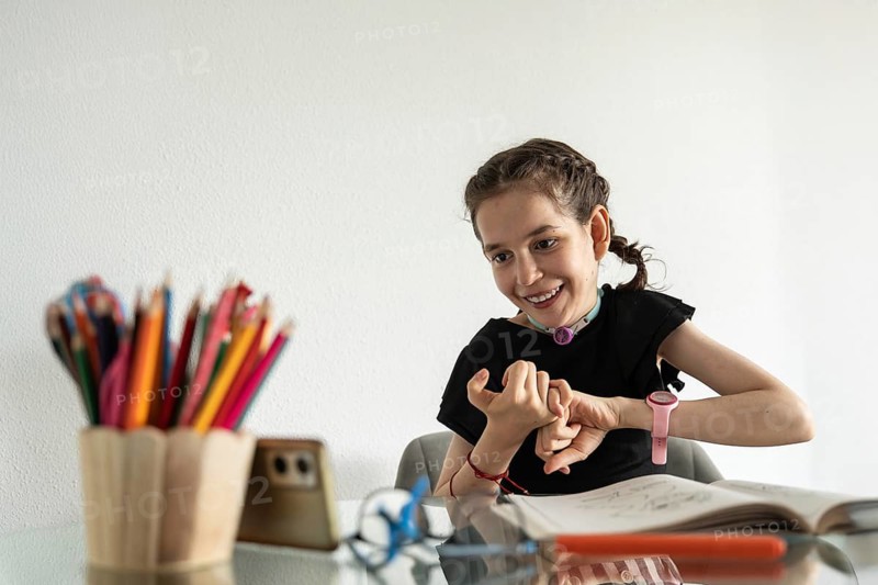 A young girl with a tracheostomy sits at a desk, surrounded by colorful pencils and a sketchbook, smiling and focused on her creative drawing while watching tutorial on phone