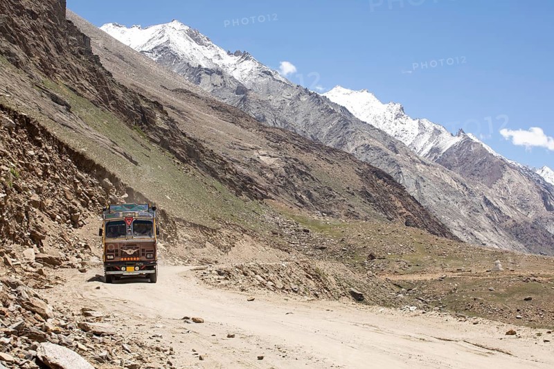 A colorful truck traverses a rugged dirt road in the stark, mountainous landscape of Zanskar Valley, Ladakh, India, with snow capped peaks under a clear blue sky