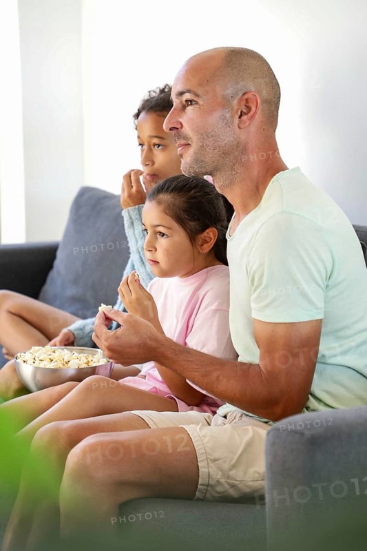 A family of three sits on a cozy sofa, engrossed in a movie while sharing a bowl of popcorn. The atmosphere is relaxed and homely, emphasizing quality family time