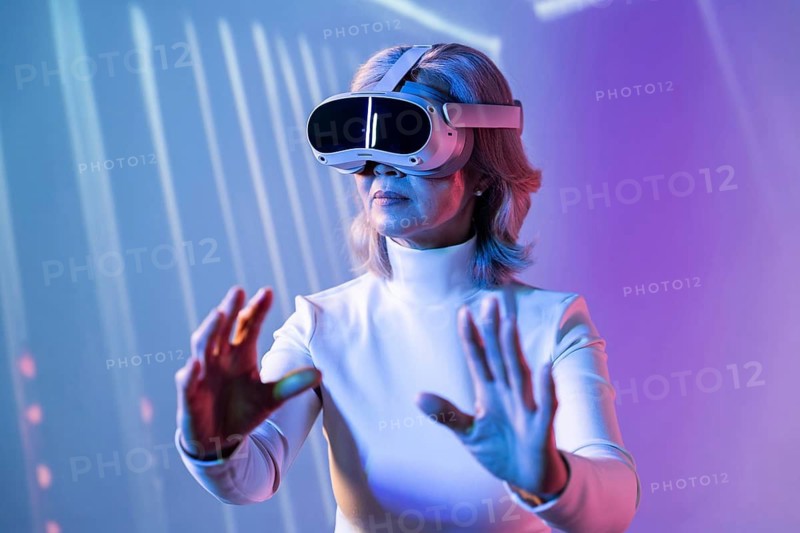 An elderly Asian woman wearing a virtual reality headset, interacting with a digital environment. The colorful background suggests an immersive metaverse experience