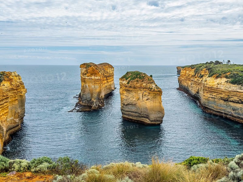 Awe inspiring coastal rock formations create a seascape as they rise from the ocean beneath a cloudy sky in the Twelve Apostles, Australia. The lush greenery enhances the scene natural beauty