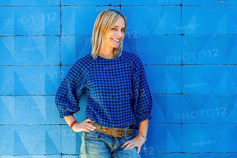 A cheerful woman in a blue checkered shirt poses casually with her hand on her hip against a vibrant blue, textured wall, looking at camera
