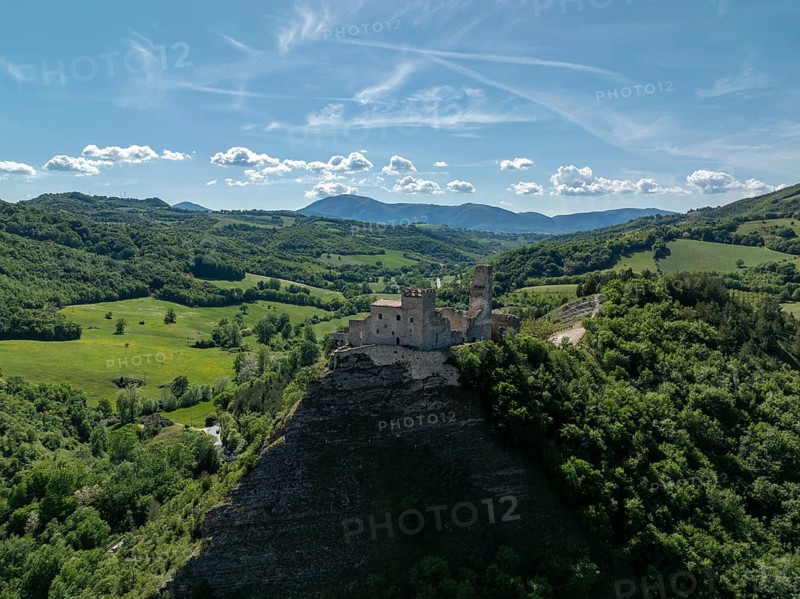 Drone view of a historic stone castle perched on a rocky hill surrounded by green rolling hills and valleys in Marche, Italy. A clear sky and distant mountains complete the scenic landscape