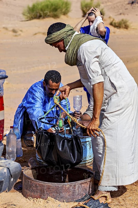 Two men in traditional dress work together to collect water in an arid landscape, Tuareg draw water from a well to wash themselves in the Sahara desert in Algeria