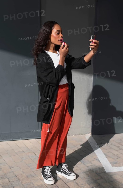 A young black woman in casual clothing applies lipstick outdoors using a compact mirror. She stands on a tiled surface, enjoying the sunny day while perfecting her look