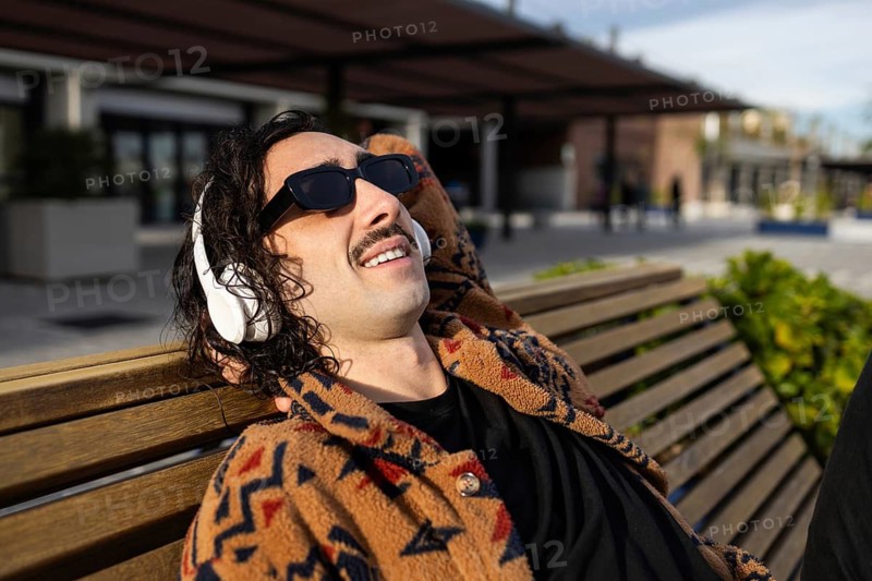 A man with a mustache lounges on a park bench, enjoying the sun He wears sunglasses and headphones, conveying a laid-back lifestyle in an urban outdoor setting