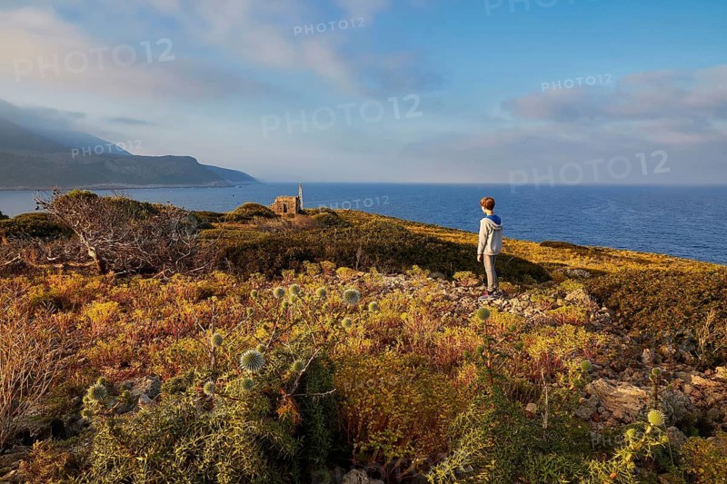 A boy stands on a coast and looks at the sea under a blue sky, morning light, Tsichli Babas islet, Navarino Bay, Pylos, Peloponnese, Greece