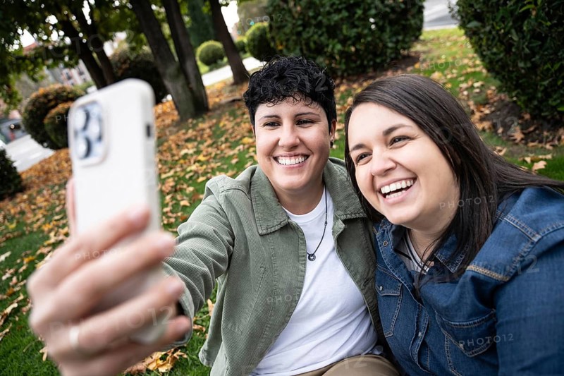 A joyful curvy lesbian couple takes a selfie in a park surrounded by autumn leaves. Their happiness and connection are palpable, capturing a moment of pure delight
