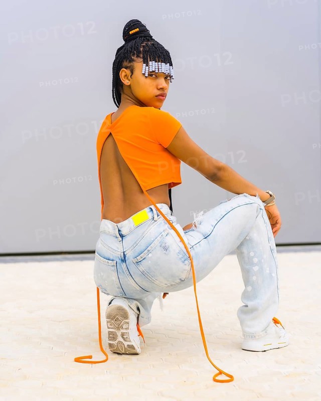 A young trap dancer with braids. Black ethnic girl with orange t-shirt and cowboy pants. Backs of Back Agachada looking seductively