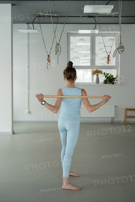 A woman in a light blue outfit practices yoga movements with a wooden stick in a spacious studio, embracing grace and discipline. Sunlight filters through the window
