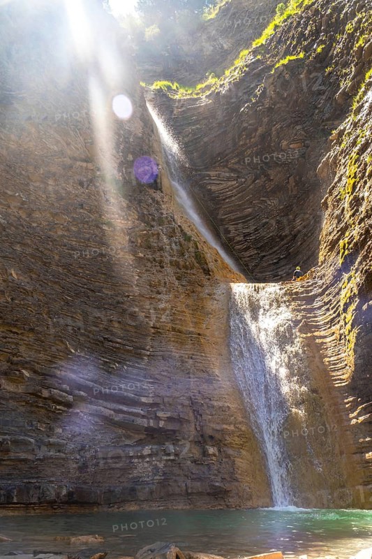 Sunlight filters through a majestic waterfall cascading down a rocky cliff face in the pyrenees mountains, creating a breathtaking natural spectacle