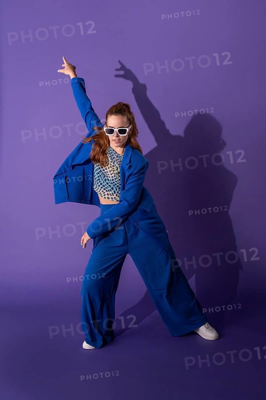 A young woman in a vibrant outfit dances energetically against a purple background. Her dynamic pose and expressive movement capture the essence of contemporary dance