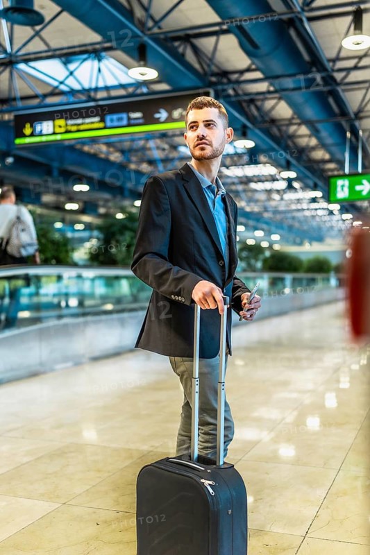 Businessman holding smartphone and pulling trolley suitcase while walking in airport terminal and looking for his gate
