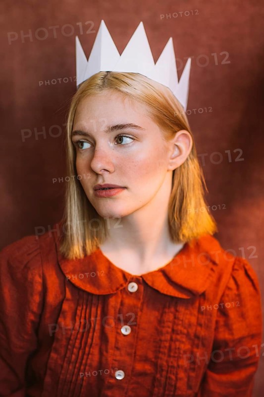 Portrait of a thoughtful teenager wearing a white paper crown. The subject is dressed in vintage red clothing, set against a warm, textured background
