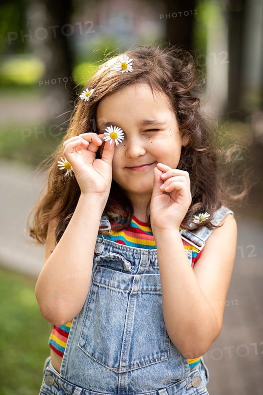 A cheerful, smiling little girl plays outdoors, holding a chamomile to her eye. Surrounded by nature, she embodies innocence and joy in a sunlit meadow setting