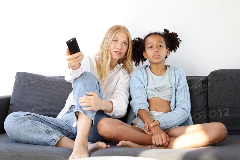 A young woman and a young girl sit on the couch holding a TV remote, watching a show together. The relaxed atmosphere highlights family bonding, shared moments, and cozy indoor leisure time