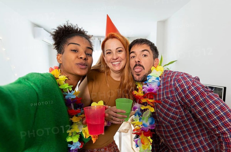 Three friends enjoying a lively birthday celebration at home. They wear colorful leis and festive outfits, holding drinks, and capturing joyful moments together