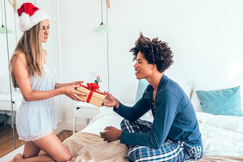 A joyful scene of a couple exchanging a wrapped gift in a cozy bedroom. The woman wears a Santa hat, symbolizing festive cheer and holiday spirit