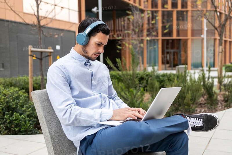 Man in business casual attire sits outside in a modern urban setting, working on his laptop while wearing headphones. His focused expression conveys productivity, remote work, and digital connectivity