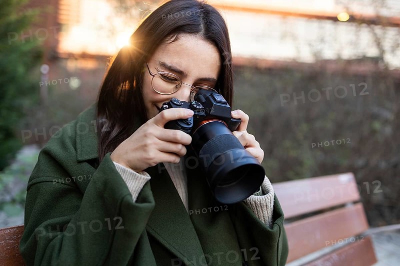 A woman wearing a green jacket is sitting on a park bench, smiling as she takes photos with a professional camera Sunlight glows softly in the background