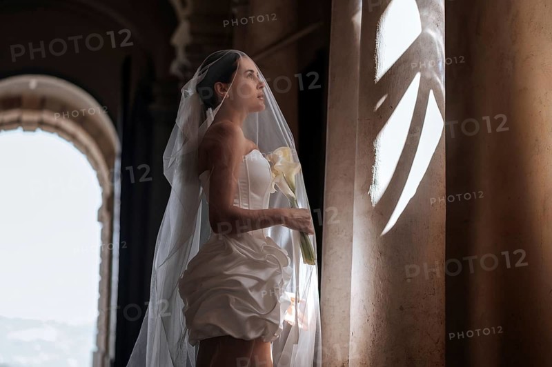 A bride in a sophisticated wedding dress stands gracefully inside an ancient Italian villa in Treia, Macerata. Soft sunlight filters through, highlighting her serene expression
