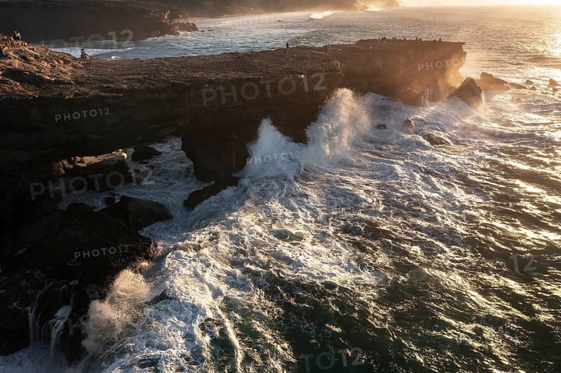 A breathtaking aerial view of the powerful waves crashing against the rocky cliffs of Riscos La Pared, Fuerteventura at sunset, with people observing nature's force