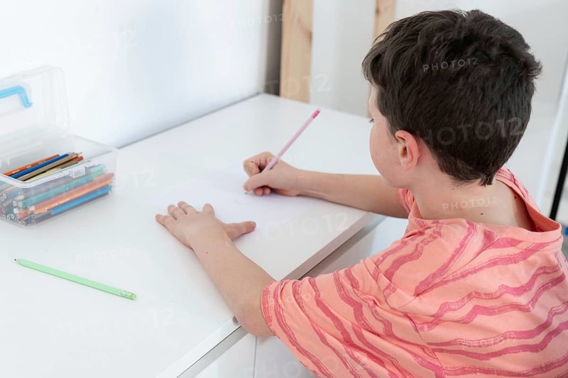 A young boy focused on drawing at a white table, surrounded by colorful pencils and school supplies in a bright, neat setting