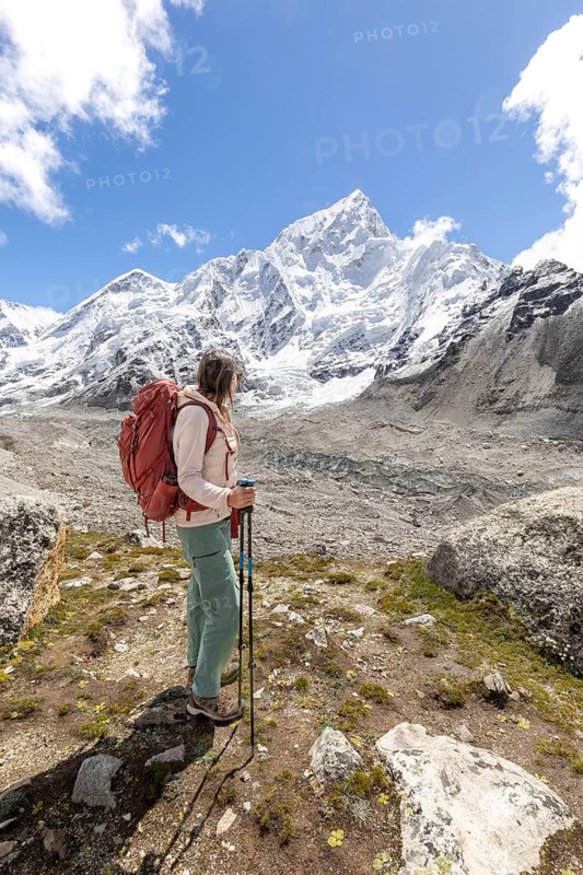A woman trekker with a backpack and trekking poles takes in the breathtaking view of the snowy Everest mountains under a clear blue sky, showcasing adventure and nature's beauty