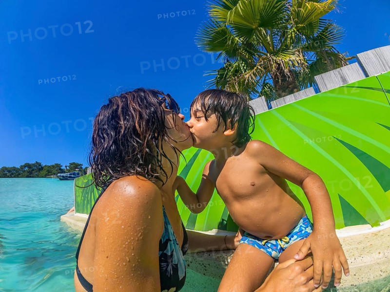 Mother and young son enjoying a tropical summer vacation, showing affection and bond while playing in a resort swimming pool with clear blue water and sunny sky