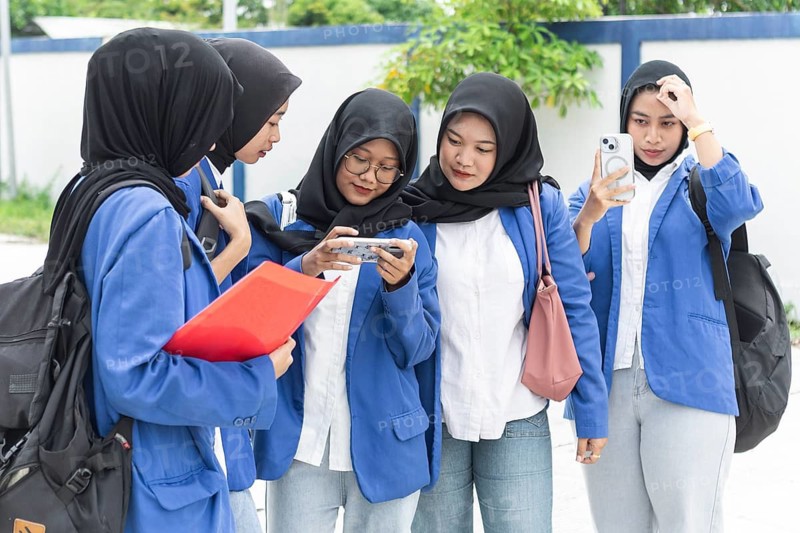 A group of Asian Muslim students wearing hijabs and blue uniforms gather outdoors. They are engaged with smartphones, illustrating technology's role in modern education