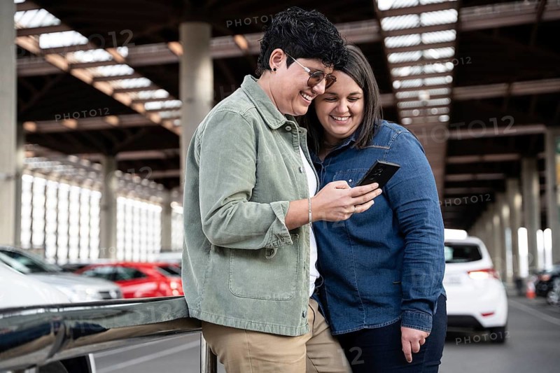 A joyful, curvy lesbian couple sharing a moment, smiling at a smartphone in a parking area. They are comfortably dressed and radiate happiness and companionship