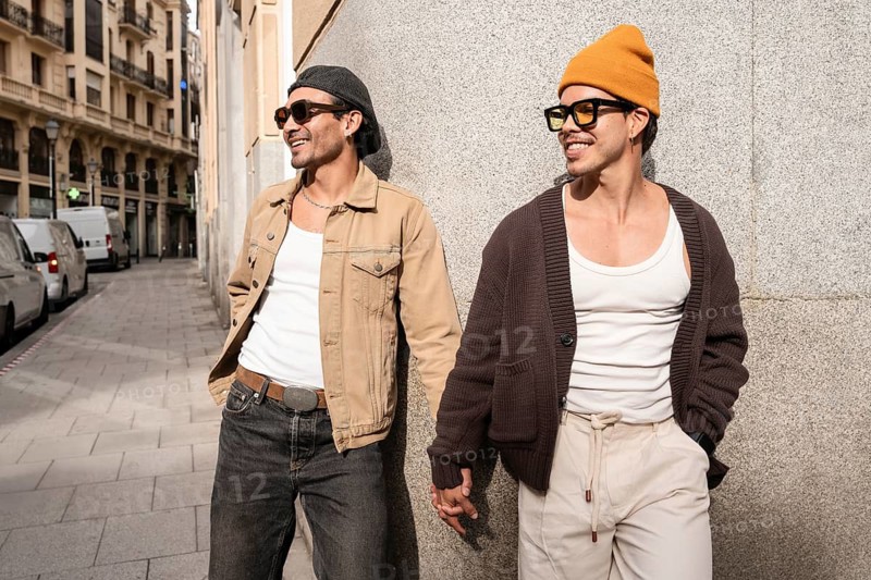 A joyful Colombian couple holding hands warmly on a city street, wearing trendy sunglasses and casual clothes, capturing a moment of affection and companionship against a brick wall backdrop