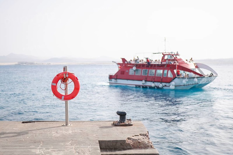The image captures a vivid port scene with a bright red ferry approaching In the foreground, a vibrant lifebuoy is mounted on the dock, enhancing maritime safety