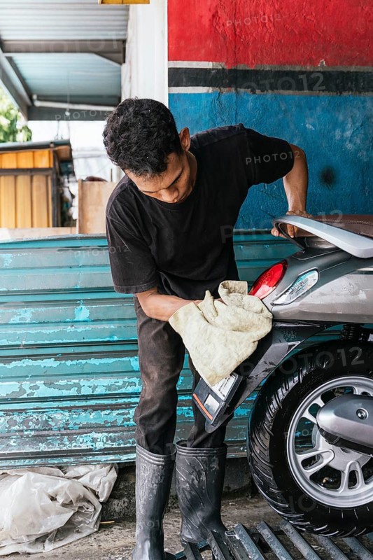 A motorcycle cleaner is meticulously polishing a bike's rear at a workshop. The scene captures dedication and care in maintaining the vehicle's pristine appearance