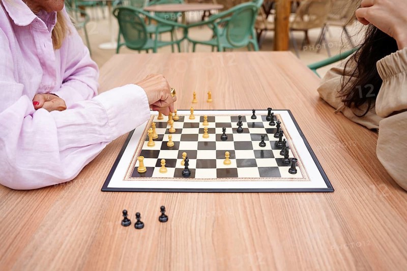 Mother and daughter engage in a chess game outside on a wooden table The focus is on the strategic move of the player in pink Green chairs and casual ambiance surround them