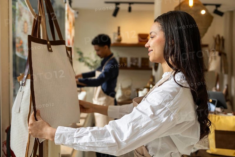 A woman examines a stylish bag in a well lit store, with a background man arranging items, showcasing a shopping scene filled with elegant bags and attentive service