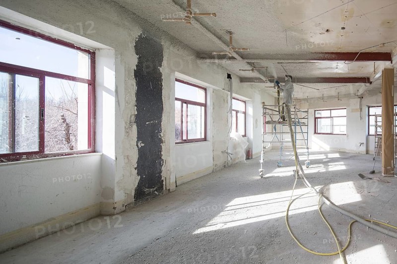 A spacious, well lit room under renovation, featuring scaffolding and a worker in protective gear spraying the ceiling. Sunlight pours through large windows