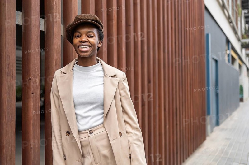 A stylish black woman smiles confidently while leaning against a wooden fence in a modern urban setting She exudes charm and elegance in casual attire