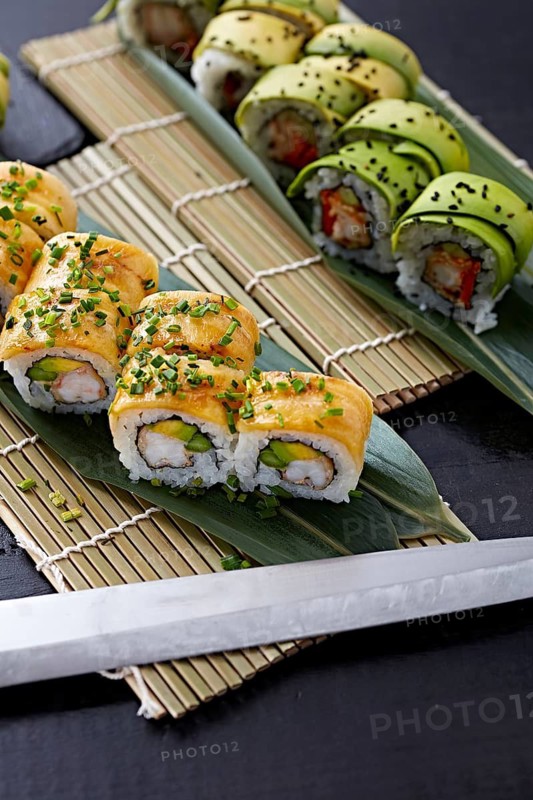 Variety of sushi rolls elegantly placed on a bamboo mat, with avocado and seafood toppings, garnished with sesame seeds and green onions