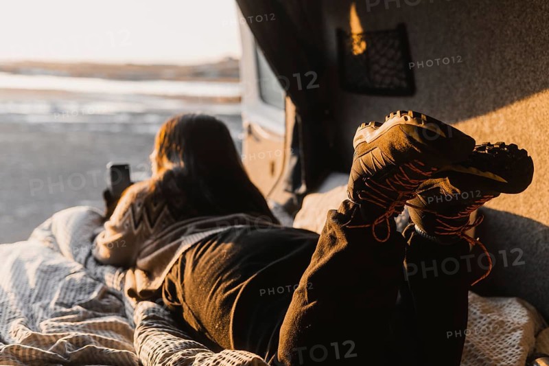 Back view of unrecognizable Latina woman, likely Colombian, lying in a camper van and using a smartphone. Captured at Camp Iceland during sunset, highlighting the serene outdoors and cozy interior setup