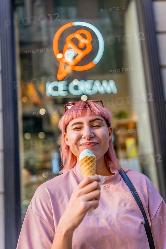 Vertical photo of a young woman with pink colored hair wink to the camera eating ice cream outdoors