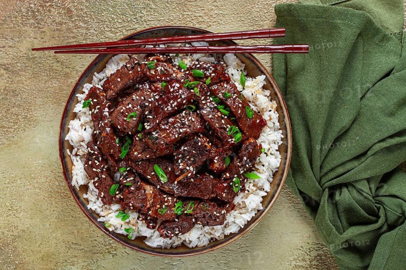 Crispy beef, with green onions and sesame seeds, on a rice pad, in a bowl with chopsticks, on a textured surface, no people