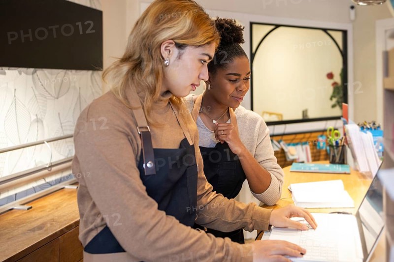 Two cosmetics store employees working together, focused on a laptop screen. They are wearing aprons, surrounded by office supplies, demonstrating teamwork and collaboration
