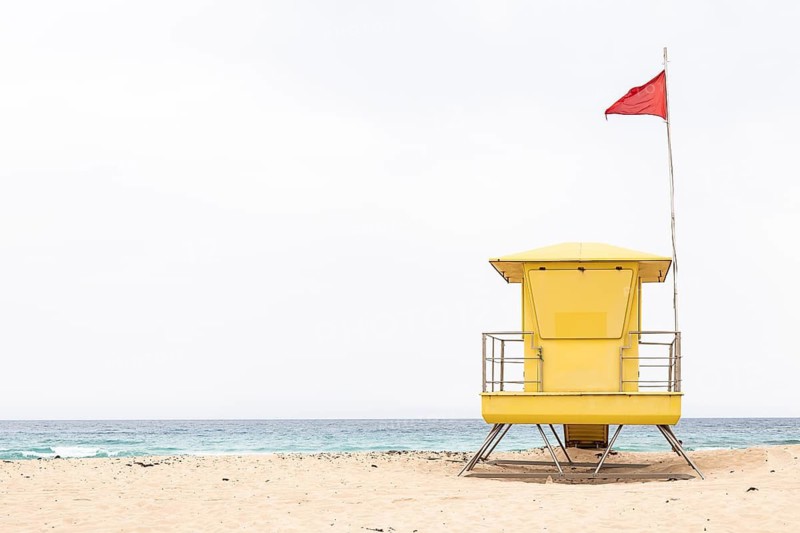 A vivid yellow lifeguard tower stands boldly on the sandy shores of Fuerteventura, Canary Islands, with a red flag fluttering against a stark backdrop of a tranquil ocean and clear sky