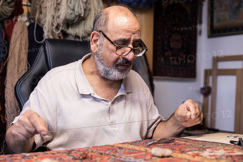An Iranian skilled artisan meticulously restores a carpet in his Salzburg workshop, surrounded by tools of his craft
