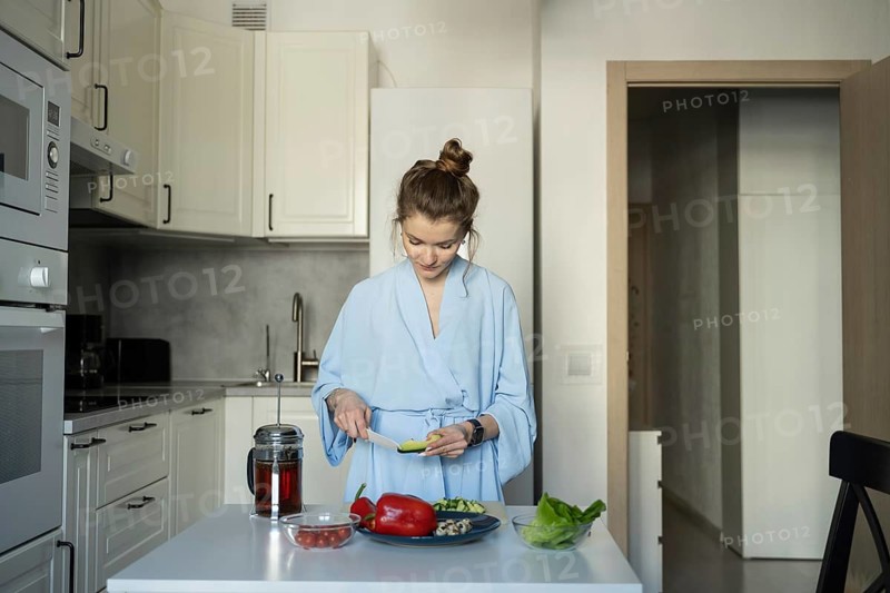 A woman in a blue robe, slicing vegetables for a healthy meal in her modern kitchen. Fresh ingredients and a peaceful atmosphere highlight a relaxing home routine