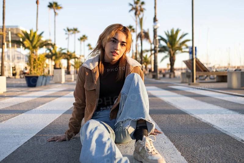 A casually dressed Mixed-race woman sits on a pedestrian crossing on a boardwalk, surrounded by palm trees and soft evening light, creating a serene and relaxed atmosphere