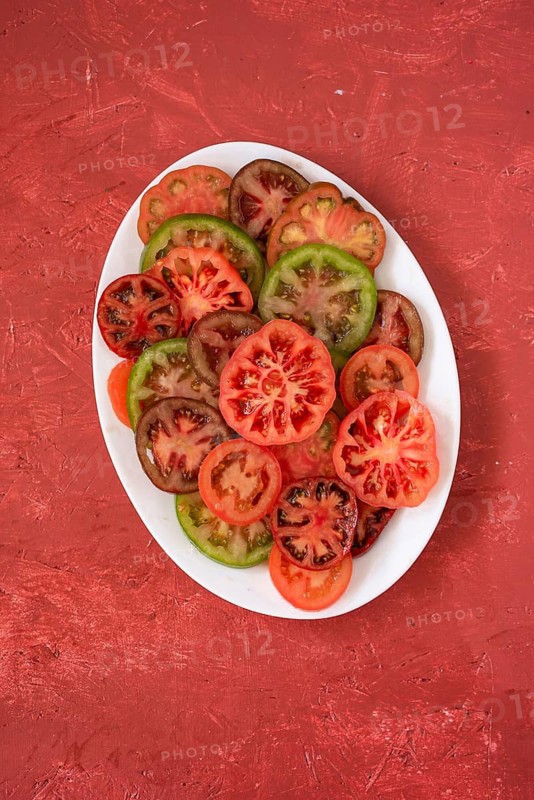 A variety of fresh heirloom tomatoes, sliced and arranged on a white plate, showcasing red, green, and brown hues. The plate sits on a textured red surface, enhancing the vibrant colors